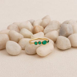 Puede incluir: Un anillo de oro con cuatro esmeraldas ovaladas. El anillo se exhibe sobre piedras blancas, contrastando las brillantes gemas verdes con el fondo neutro. La banda del anillo es delgada y delicada.