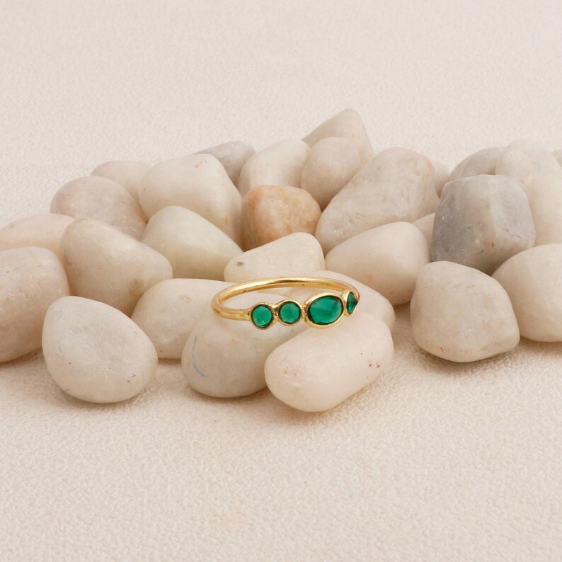 Puede incluir: Un anillo de oro con cuatro esmeraldas ovaladas. El anillo se exhibe sobre piedras blancas, contrastando las brillantes gemas verdes con el fondo neutro. La banda del anillo es delgada y delicada.