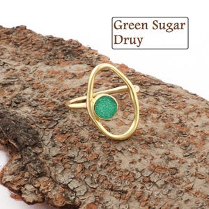 Può includere: Un anello color oro con una pietra verde e circolare al centro di un design ovale. L'anello è presentato su un pezzo di legno strutturato. Il testo "Green Sugar Druy" è in un riquadro bianco.