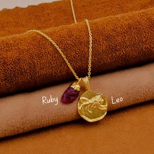 以下が含まれることがあります： 2つのペンダントが付いたゴールドカラーのネックレス。1つは生のルビーの宝石、もう1つは獅子座のデザインの円形ペンダントです。「Ruby」と「Leo」の文字が書かれています。ネックレスは茶色の表面に表示されています。