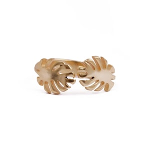 May include: A gold-colored ring featuring a split design with two stylized palm leaf motifs. The ring has a smooth, matte finish and is set against a white background. The ring's design is symmetrical, with each leaf mirroring the other.