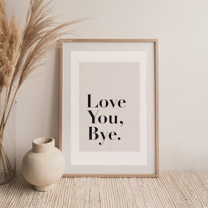 May include: A framed print with a light pink background and the text "Love You, Bye." in black lettering. The print is sitting on a light brown woven rug in front of a white wall. A vase with dried pampas grass is on the left side of the print and a small beige ceramic vase is on the right.