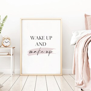 May include: A gold framed print with the text "WAKE UP AND make up" in black font with a pink watercolor-style brushstroke behind the word "make".