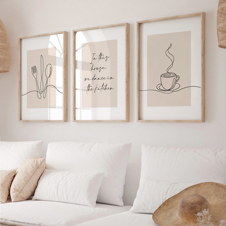 Peut inclure: Trois impressions d'art encadr&eacute;es avec des dessins au trait minimalistes. L'une repr&eacute;sente des couverts, une autre porte le texte "In this house, we dance in the kitchen", et la troisi&egrave;me repr&eacute;sente une tasse &agrave; caf&eacute;. Les impressions ont une palette de couleurs neutres.