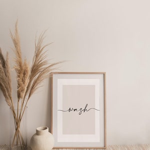 May include: A framed print with a light beige background and the word "wash" written in black cursive script. The print is sitting on a woven rug in front of a vase with dried pampas grass.