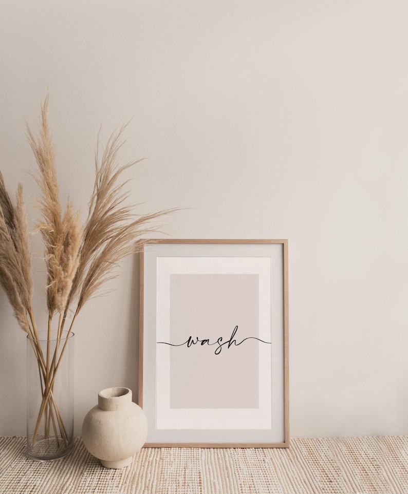 May include: A framed print with a light beige background and the word "wash" written in black cursive script. The print is sitting on a woven rug in front of a vase with dried pampas grass.