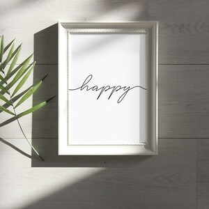 May include: A white frame with a white mat and a black cursive word "happy" on a white background. The frame is on a light gray wood surface with a green leaf shadow.