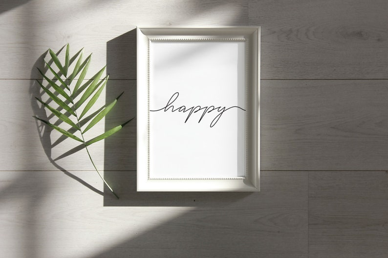 May include: A white frame with a white mat and a black cursive word "happy" on a white background. The frame is on a light gray wood surface with a green leaf shadow.