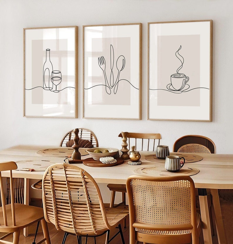 May include: Three minimalist line art prints in wooden frames, featuring a wine bottle and glass, a fork, knife, and spoon, and a steaming cup of coffee. The prints are hung on a white wall above a wooden dining table with wicker chairs.