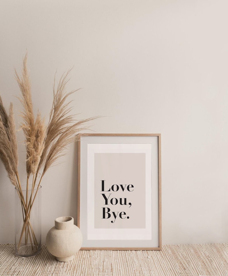 May include: A framed print with a light pink background and the text "Love You, Bye." in black lettering. The print is sitting on a light brown woven rug in front of a white wall. A vase with dried pampas grass is on the left side of the print and a small beige ceramic vase is on the right.