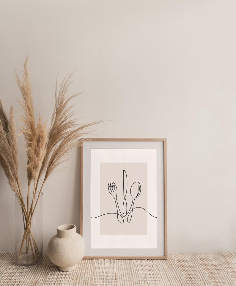 May include: A framed print featuring a minimalist line drawing of a fork, knife, and spoon on a light brown background. The print is in a wooden frame and is resting on a woven rug.