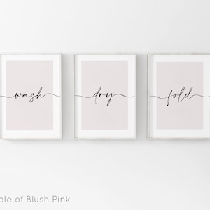 May include: Three framed prints with a blush pink background and black calligraphy. The prints read "wash", "dry", and "fold" in a cursive font.