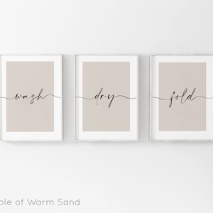 May include: Three framed prints with a light brown background and white frames. The prints have the words "wash", "dry", and "fold" written in black cursive.