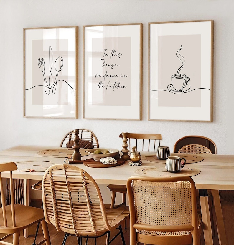 Peut inclure: Trois impressions encadr&eacute;es avec des dessins au trait minimalistes. L'une repr&eacute;sente des couverts, une autre dit "In this house, we dance in the kitchen", et la troisi&egrave;me montre une tasse &agrave; caf&eacute;. Les impressions sont dans des cadres en bois, avec une palette de couleurs neutres.