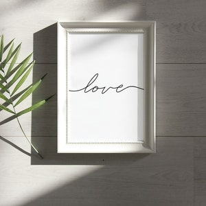 May include: A white framed print with the word "love" written in black cursive script. The print is on a white wooden surface with a green leaf.