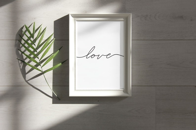 May include: A white framed print with the word "love" written in black cursive script. The print is on a white wooden surface with a green leaf.