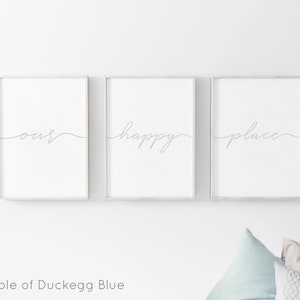 May include: Three white framed prints with the words "our", "happy", and "place" written in a cursive script in a light blue color. The prints are hung on a white wall.