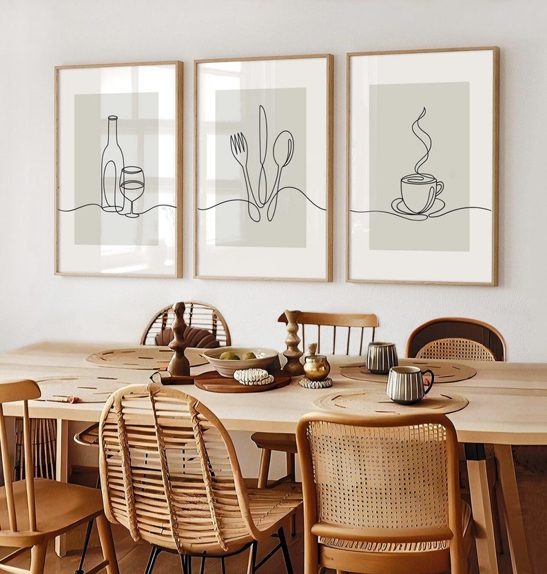 May include: Three minimalist line art prints in wooden frames, hanging on a white wall above a wooden dining table with wicker chairs. The prints depict a bottle of wine and a glass, a fork, knife, and spoon, and a steaming cup of coffee.