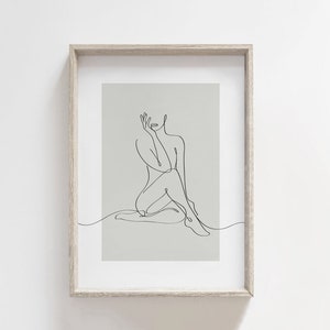 May include: A black and white line drawing of a person sitting with their leg crossed. The drawing is on a light gray background and is framed in a light wood frame.