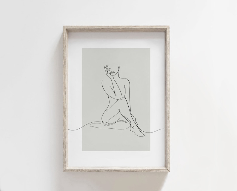 May include: A black and white line drawing of a person sitting with their leg crossed. The drawing is on a light gray background and is framed in a light wood frame.