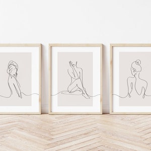 May include: Set of three minimalist line art prints featuring female figures in various poses. The prints are framed in light wood frames and have a beige background.