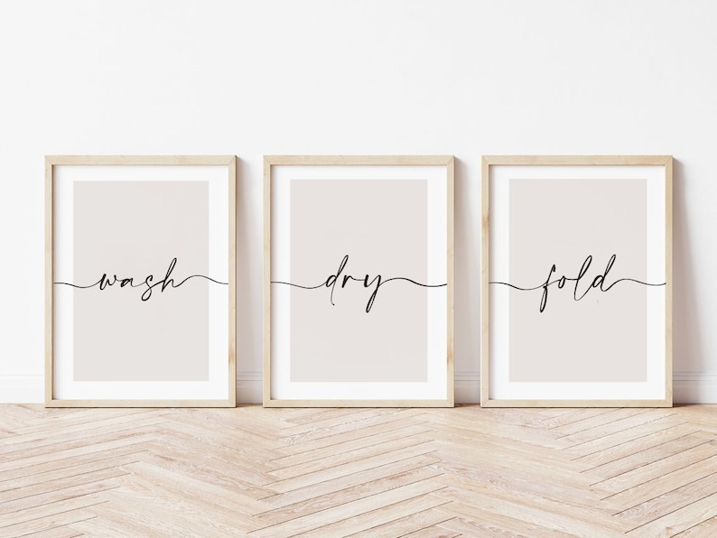 May include: Three framed prints with a light beige background and black calligraphy. The prints read "wash", "dry", and "fold" in a cursive font.