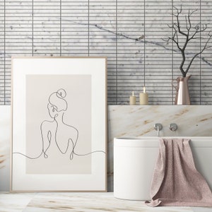 May include: A framed line drawing of a woman with her hair in a bun, hanging on a white tiled wall above a white bathtub with a pink towel draped over the side.