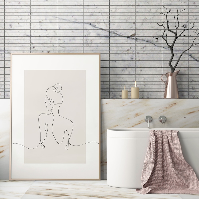 May include: A framed line drawing of a woman with her hair in a bun, hanging on a white tiled wall above a white bathtub with a pink towel draped over the side.