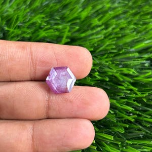 Puede incluir: Una pequeña gema hexagonal facetada con un tono púrpura claro. La gema se sostiene en una mano sobre un fondo de césped artificial verde. Las facetas de la gema capturan la luz, creando un efecto brillante.