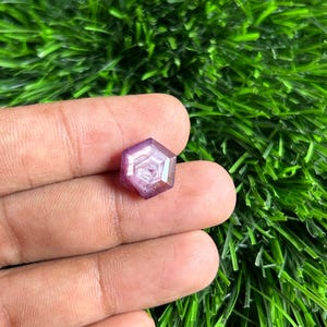 Puede incluir: Una gema hexagonal facetada con un tono púrpura. La gema tiene un patrón interno complejo y se sostiene en una mano sobre un fondo de césped artificial verde. Las facetas de la gema capturan la luz, creando un efecto brillante.