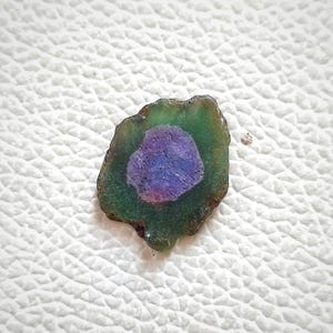 Puede incluir: Una piedra preciosa verde y morada con una forma irregular y rugosa. La piedra preciosa tiene un centro circular morado rodeado por una capa exterior verde.