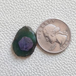 Könnte beinhalten: Ein grüner und violetter Edelstein mit einer rauen, unregelmäßigen Form. Der Edelstein ist etwa so groß wie eine US-amerikanische 25-Cent-Münze.