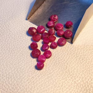 May include: A collection of small, round, faceted ruby gemstones in various shades of pink and red. The gemstones are scattered on a white surface.
