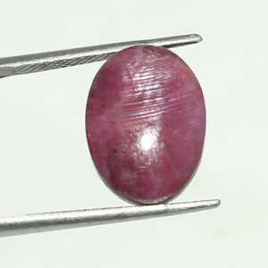May include: A polished, oval gemstone in deep red and pink hues, held by silver tweezers. The gem's surface is smooth with faint linear patterns. The background is plain white.