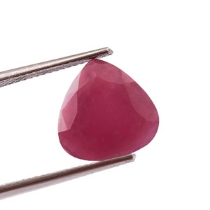 Pode incluir: Uma gema rosa em forma de pêra segurada por pinças. A gema tem um corte facetado e é translúcida.