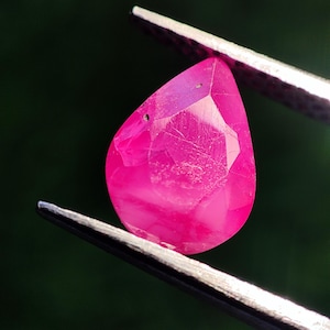 Könnte beinhalten: Ein facettierter, tropfenförmiger Rubin-Edelstein, gehalten von einer Pinzette. Der Edelstein hat eine leuchtende rosa-rote Farbe, mit sichtbaren inneren Einschlüssen und einer polierten Oberfläche. Der Hintergrund ist ein verschwommenes, dunkles Grün.