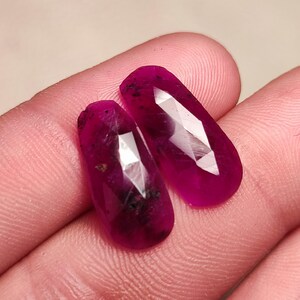 May include: Two faceted, oval-shaped gemstones with a deep pink hue. The stones have a slightly darker, almost black, inclusion in the centre.
