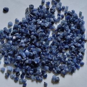 以下が含まれることがあります： 小さな粗い青いサファイアの宝石が大量に積み重ねられています。宝石はさまざまな青の色合いで、自然で研磨されていない外観をしています。