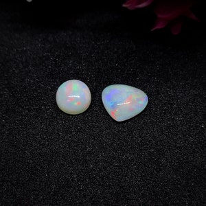 Puede incluir: Dos piedras preciosas de ópalo iridiscente, una redonda y otra en forma de lágrima, sobre un fondo negro.