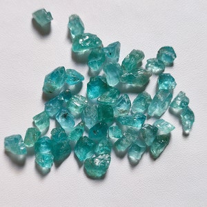 May include: A collection of teal blue gemstone roughs, with various shapes and sizes. The roughs are scattered on a white surface.
