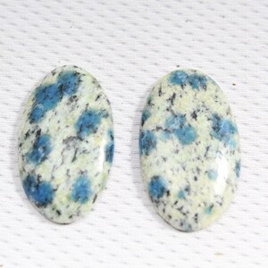 May include: Two oval-shaped blue and white jasper cabochons. The cabochons have a speckled pattern of blue and white with black veins.