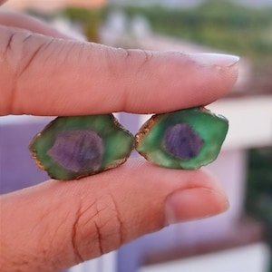 May include: Two polished gemstones, each with a green outer layer and a central purple core. The edges of the stones have a golden-brown hue. The stones are held between fingers, showcasing their natural beauty.