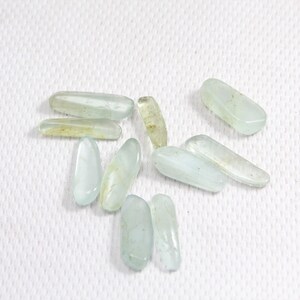 May include: A collection of ten smooth, light blue aquamarine stones, each with a slightly different shape and size. The stones are arranged on a white textured surface.