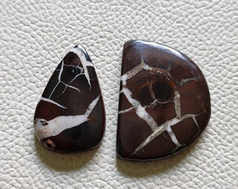 May include: Two cabochon gemstone pendants with a brown and white crackle pattern. The pendant on the left is teardrop shaped and the pendant on the right is a half-circle.