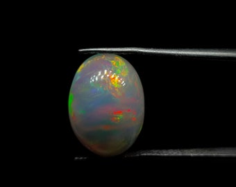 Puede incluir: Una piedra preciosa de ópalo ovalada blanca con colores arcoíris iridiscentes, sostenida por pinzas sobre un fondo negro.