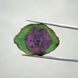 May include: A green and pink gemstone with a rough, natural shape. The gemstone is held by tweezers.