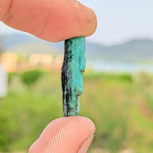 Op de afbeelding: Een ruw, langwerpig kristalexemplaar met een levendige turkoois-groene tint en diepzwarte, verticale strepen over de gehele lengte. Het mineraal heeft een ruwe, kolomvormige textuur met natuurlijke ribbels en een enigszins ongelijkmatig oppervlak.