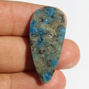 May include: A teardrop-shaped cabochon of blue and white kyanite with a speckled pattern. The stone is translucent and has a smooth, polished surface.