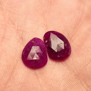 May include: Two faceted ruby gemstones, each with a teardrop shape and a deep pink hue. The stones have a faceted cut and are set in a clear, transparent material.