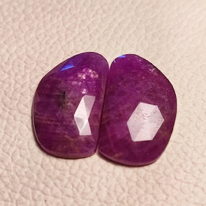 May include: Two purple gemstone cabochons with a faceted cut. The gemstones are set in a raw, natural style.